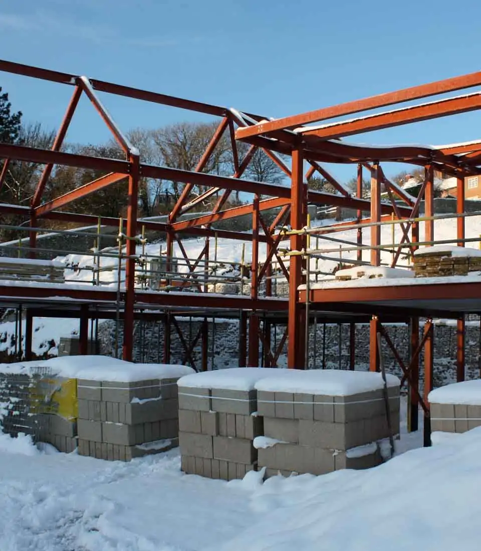 A steel framework of a building rises above snowy ground, with stacks of concrete blocks nearby, amidst a winter landscape under a clear blue sky and scattered greenery.