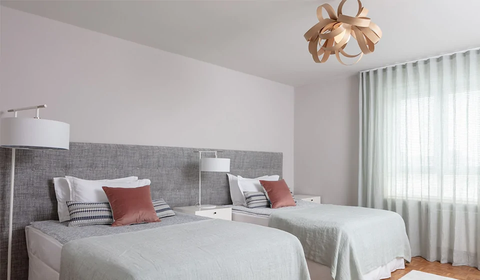 Two neatly arranged beds with light green bedspreads and decorative pillows occupy a room. A modern light fixture hangs above, and sheer curtains filter natural light from a window.