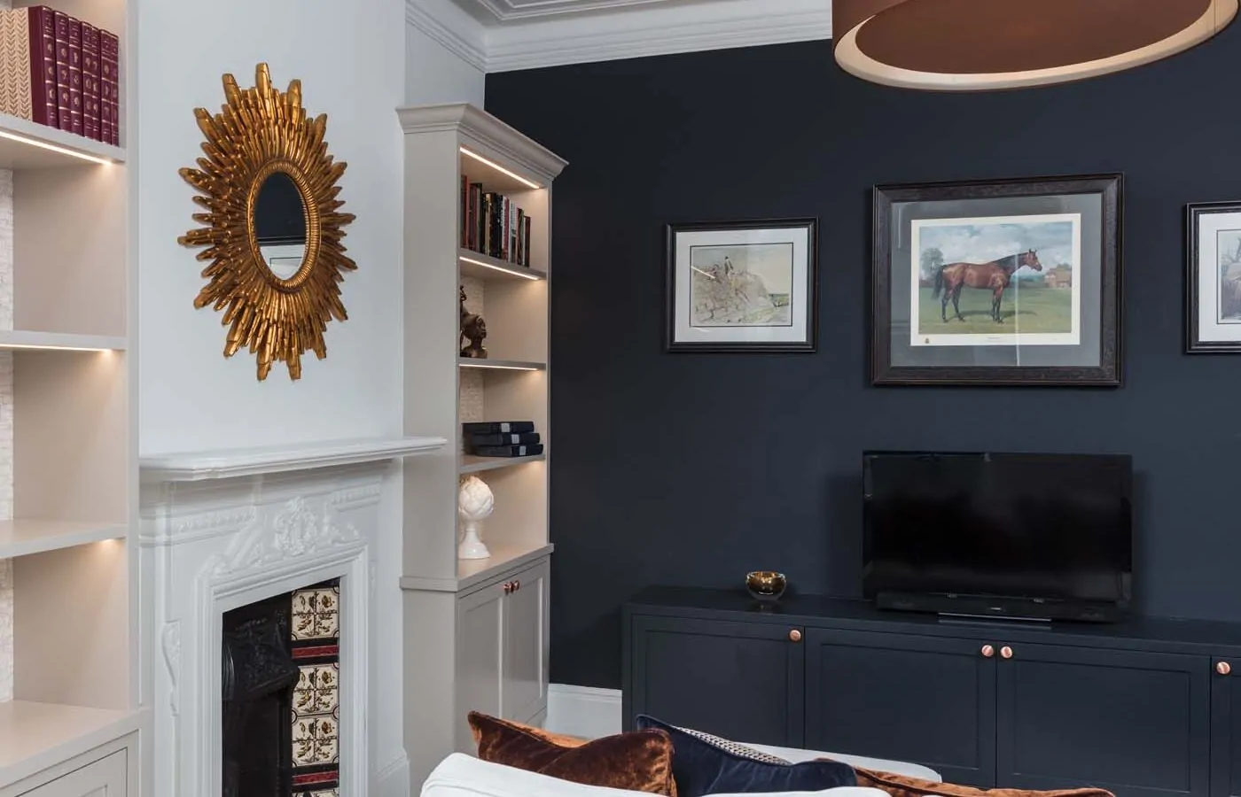 A stylish living room features a white sofa with colorful cushions, a television on a dark cabinet, and decorative shelves. A gold mirror and framed horse painting enhance the elegant ambiance.