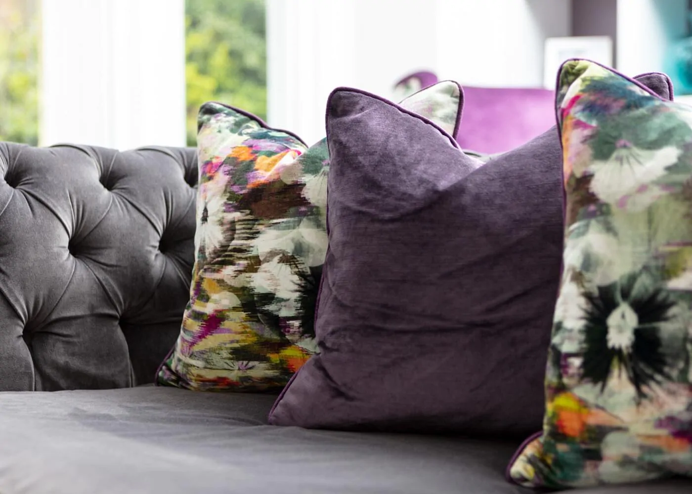 A grey tufted sofa features three decorative pillows: two are purple and one is vibrant with floral patterns, set against a light-filled modern interior.