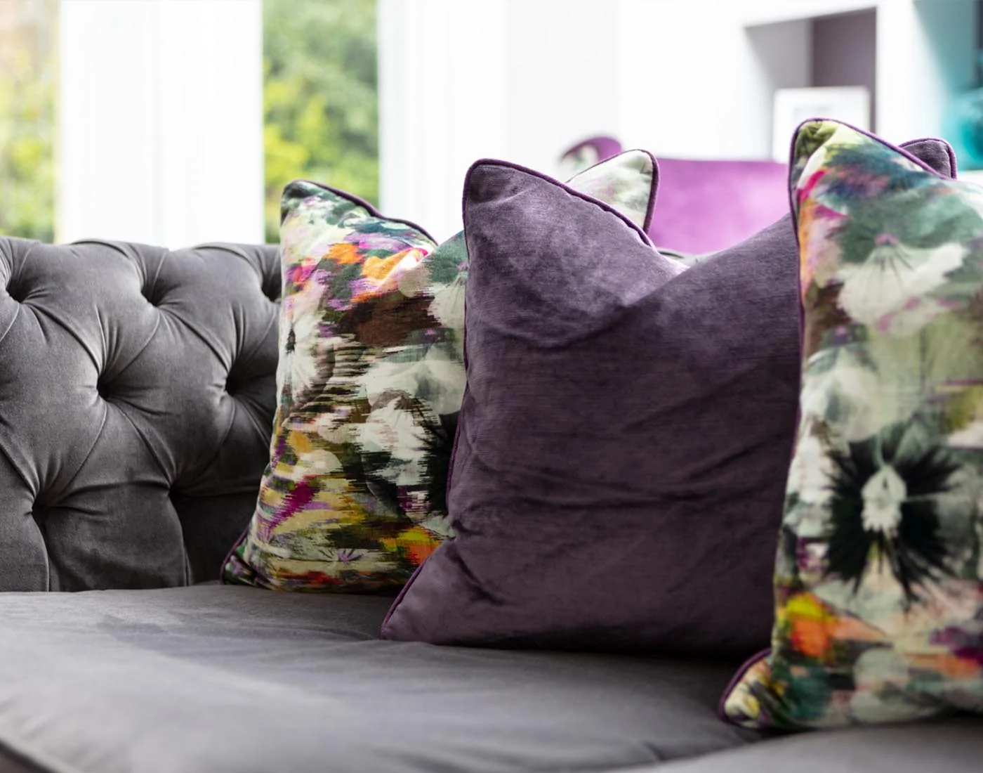 A grey tufted sofa features three decorative pillows: two are purple and one is vibrant with floral patterns, set against a light-filled modern interior.