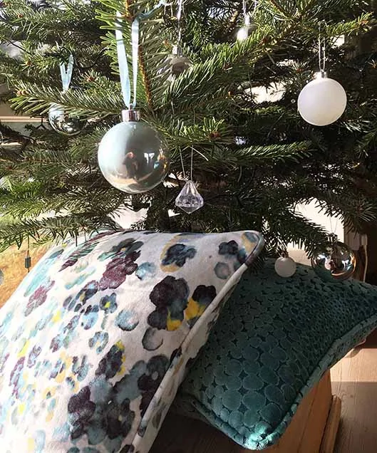 A decorated Christmas tree with hanging ornaments stands above two decorative pillows, one featuring a floral pattern and the other a textured design, placed on a wooden surface.