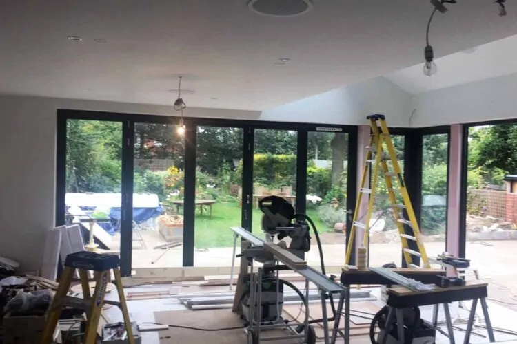 A room under renovation features construction tools, a tall ladder, and a table saw. Large glass doors open to a garden with greenery and furniture visible outside.