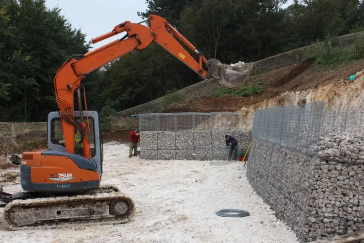 An excavator scoops gravel while two workers construct a stone wall in a gravel pit. Surrounded by trees, the site is partially cleared with visible dirt mounds and tools.