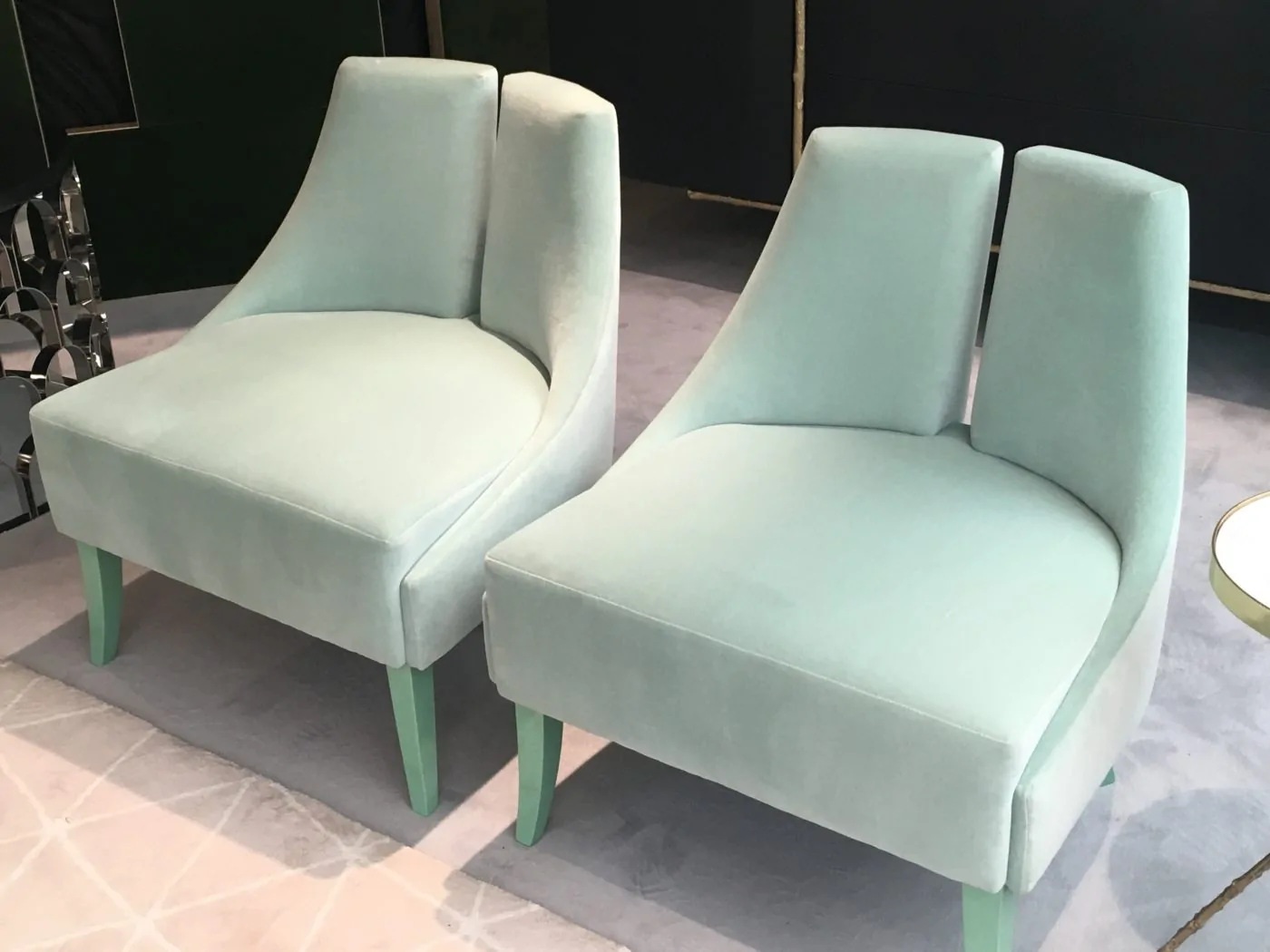 Two elegant, mint green velvet chairs sit closely together, featuring smooth curves and light green legs, on a soft, light gray patterned rug, in a stylish, contemporary setting.
