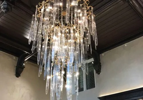 A luxurious chandelier with elongated glass pieces hangs from a dark wooden ceiling, illuminating the surrounding elegant room while contrasting with the light-colored walls.