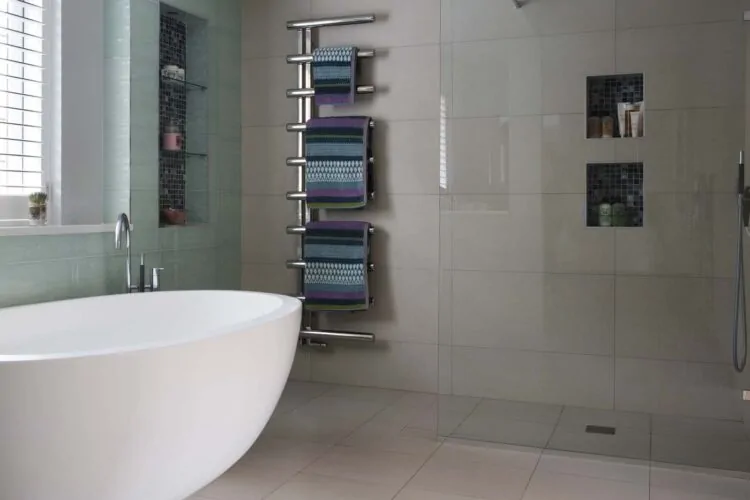 A modern bathroom features a freestanding white bathtub, a glass shower, and a towel rack with colorful towels. The walls are tiled in soft colors, creating a serene atmosphere.
