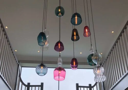 Colorful glass pendant lights hang from the ceiling, illuminating a modern staircase in a spacious interior, with large windows providing natural light and a view outside.