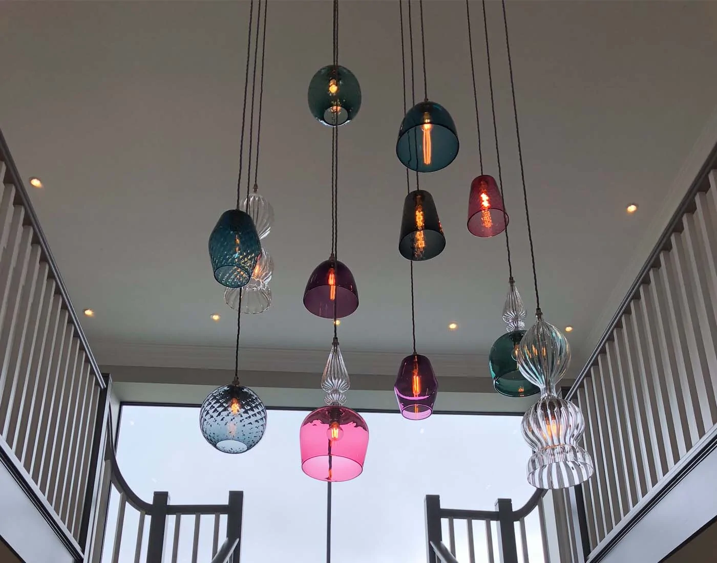 Colorful glass pendant lights hang from the ceiling, illuminating a modern staircase in a spacious interior, with large windows providing natural light and a view outside.