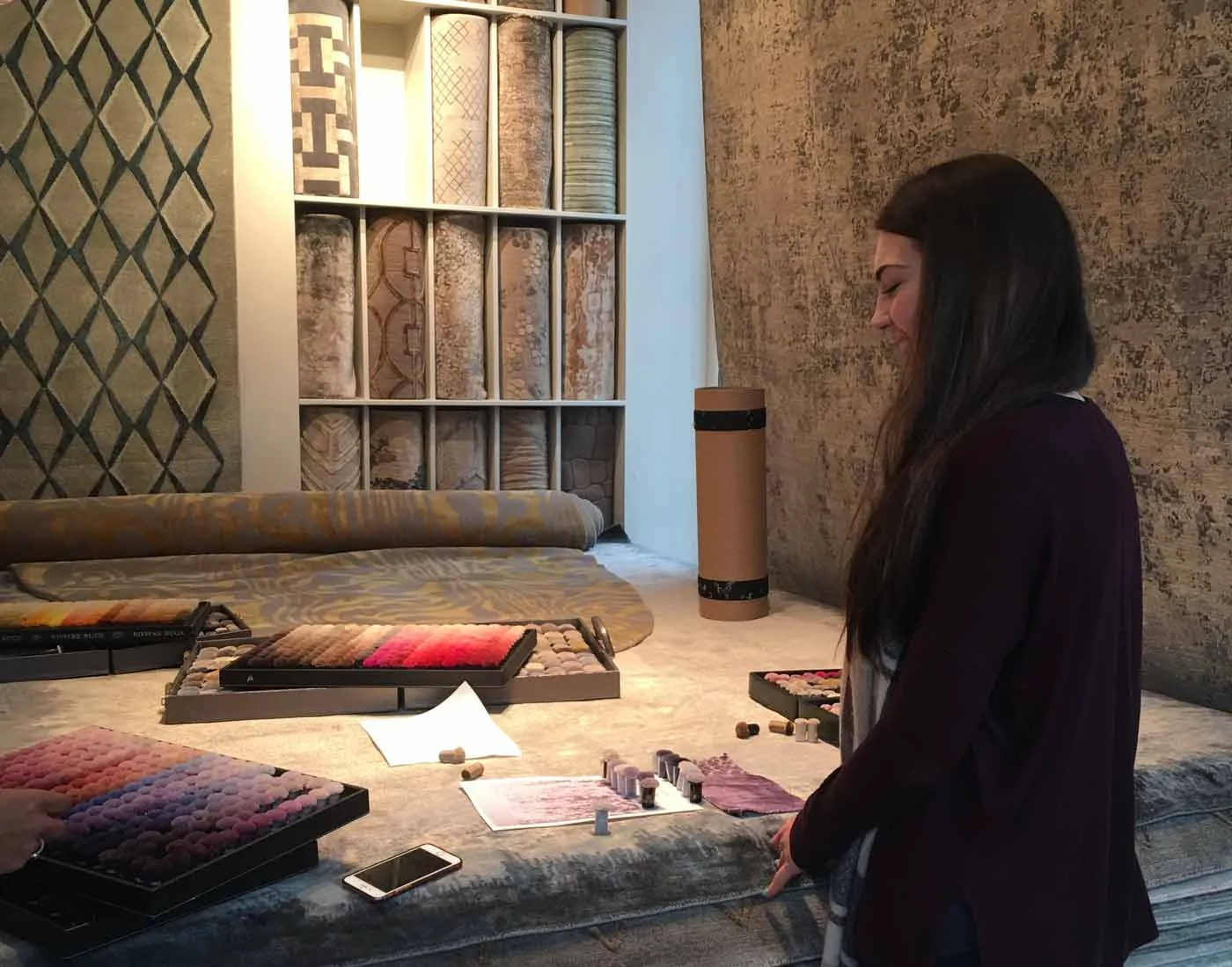 A woman examines color samples on a work table, surrounded by rolls of textured fabric and rugs on display. The environment suggests a design studio or showroom.