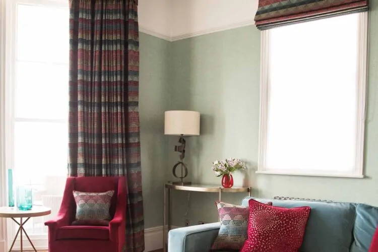 A cozy living room features a red armchair and a blue sofa adorned with colorful cushions. A side table holds blue glass vases, while a lamp and flowers complement the decor.