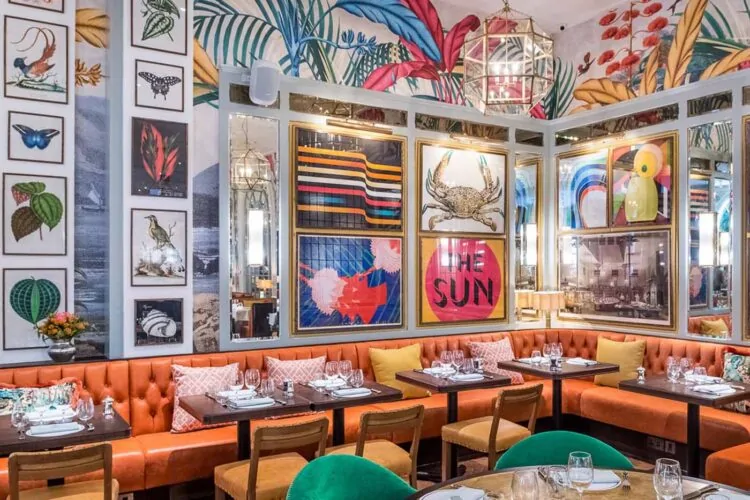 A vibrant restaurant features colorful wall art and tropical patterns. Diners sit at wooden tables with green and orange upholstery, surrounded by framed pictures and a bright, lively atmosphere.