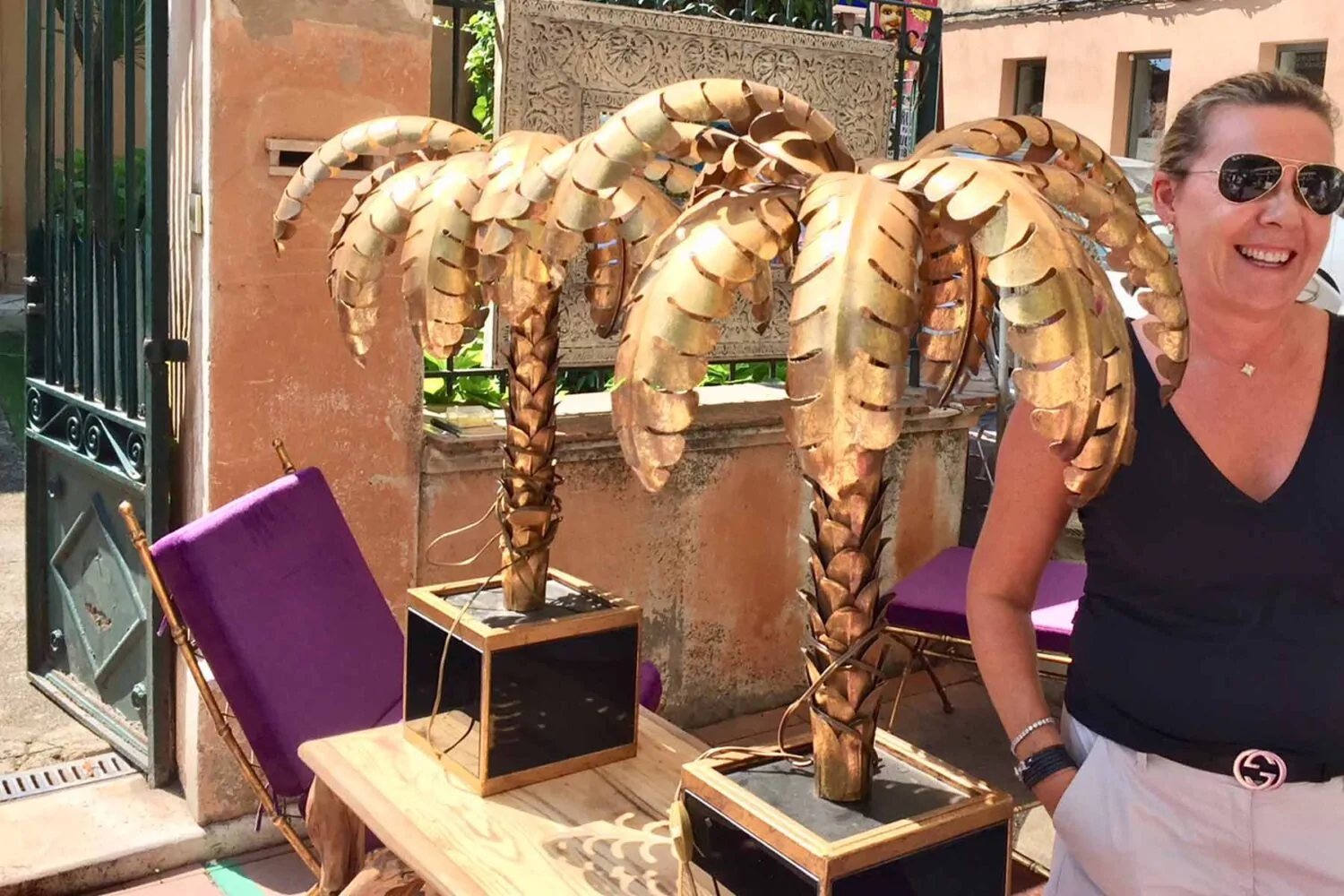Two golden palm tree sculptures stand on a table beside a smiling woman. A purple folding chair leans against the table, set against a sunny, vibrant outdoor setting.