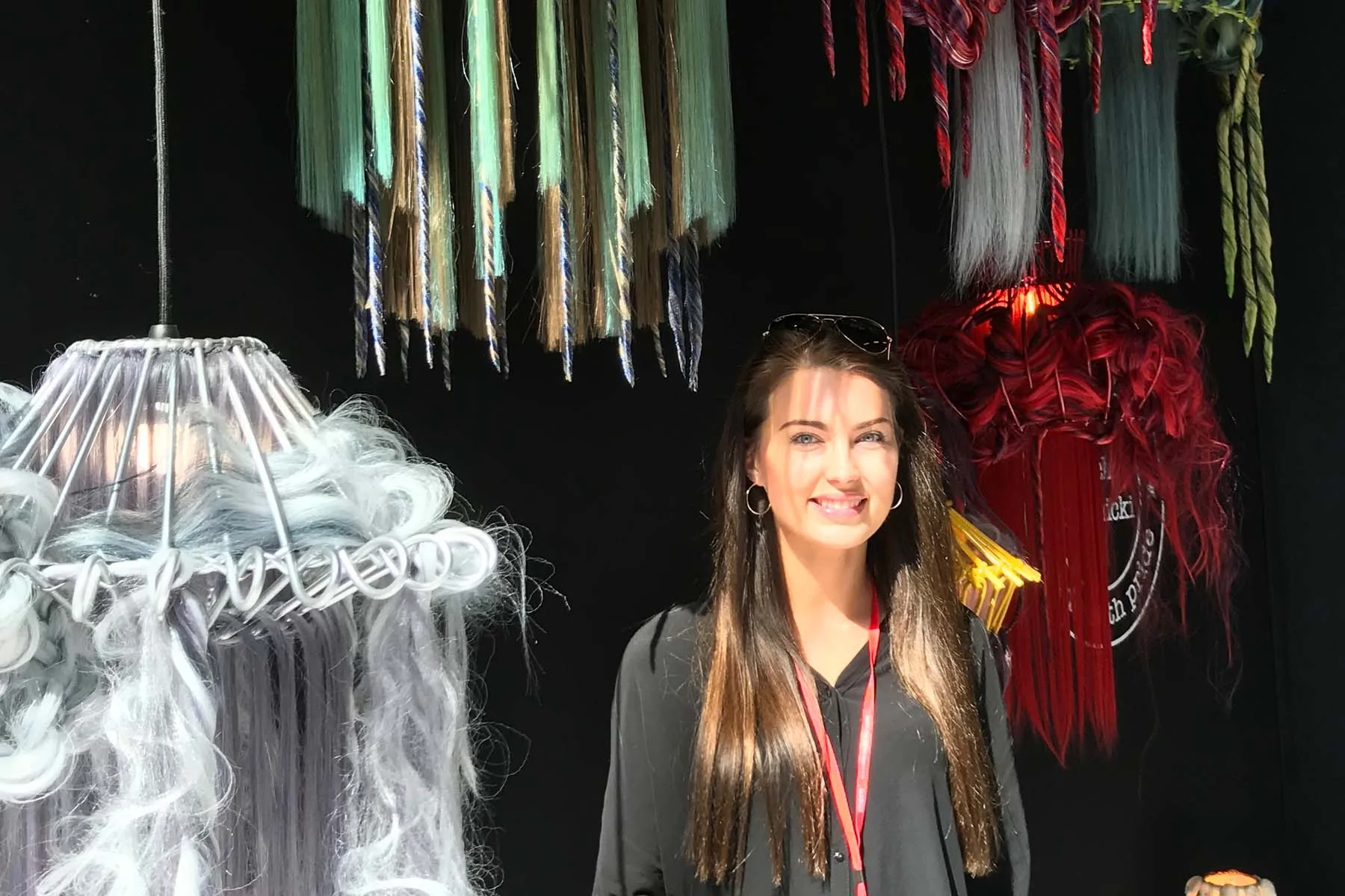 A young woman stands smiling next to a collection of colorful hanging lamps made from various materials, against a dark backdrop. Sunlight casts shadows across her face and surroundings.