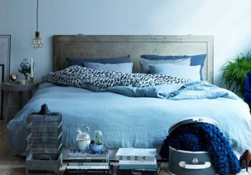 A bed adorned with blue linens and patterned pillows occupies the center. Nearby, a table holds flowers and candles. A caged decoration and stacked books add charm, while a green plant enhances the atmosphere.