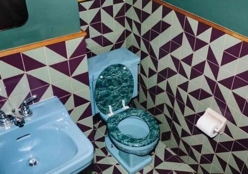 A blue sink and a toilet with a green marble seat are positioned in a bathroom surrounded by geometric patterned tiles in green and purple. A roll of toilet paper is mounted on the wall.