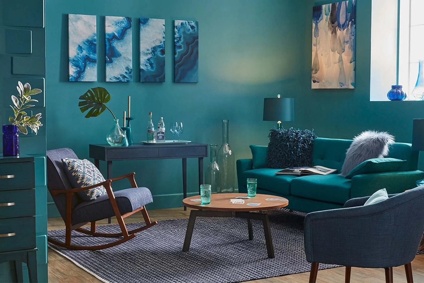 A turquoise living room features a cozy sofa with cushions and a shaggy throw, a wooden coffee table with glasses, and a rocking chair. Art and vases adorn the walls.