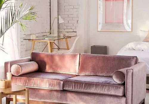 A plush pink sofa with rounded armrests is centrally placed in a bright, airy room. A desk and chair are nearby, with a plant adding a touch of greenery.