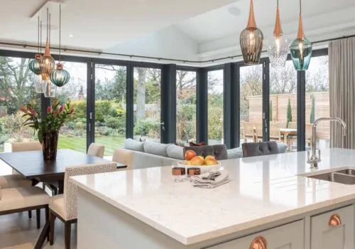A modern kitchen features a marble countertop and stylish overhead pendant lights. A dining area with a wooden table and soft chairs is visible, alongside a spacious garden view through large windows.