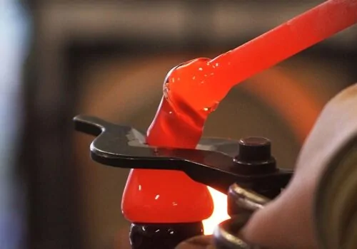 A glowing red molten glass blob is being shaped with metal tools in a workshop, surrounded by a blurred background hinting at a furnace's warm glow.