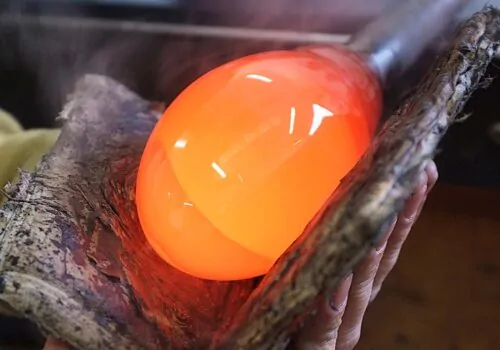 A glowing, orange glass orb rests on textured wood held by a hand, with steam rising around it, indicating a hot glassblowing process in a workshop environment.