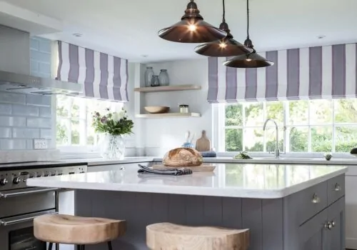 A marble countertop island with wooden stools displays a loaf of bread, while three pendant lights illuminate the modern kitchen featuring blue subway tiles and a flower vase by the window.