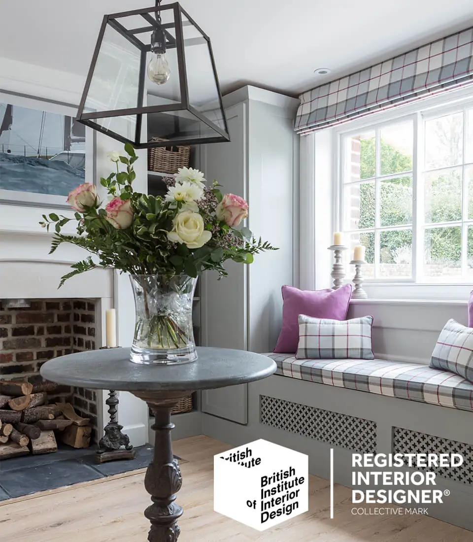 A round, stone tabletop holds a clear vase filled with assorted roses and greenery. Natural light floods a cozy room with a fireplace and a cushioned window seat, enhancing the inviting atmosphere.