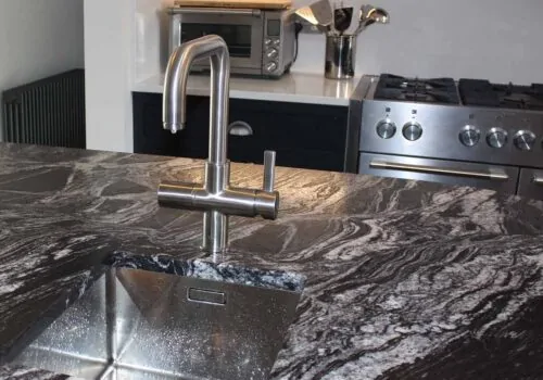 A sleek, silver kitchen faucet is positioned above a rectangular stainless steel sink, which sits on a dark, patterned countertop. In the background, a stove and microwave are visible, suggesting a modern kitchen setting.