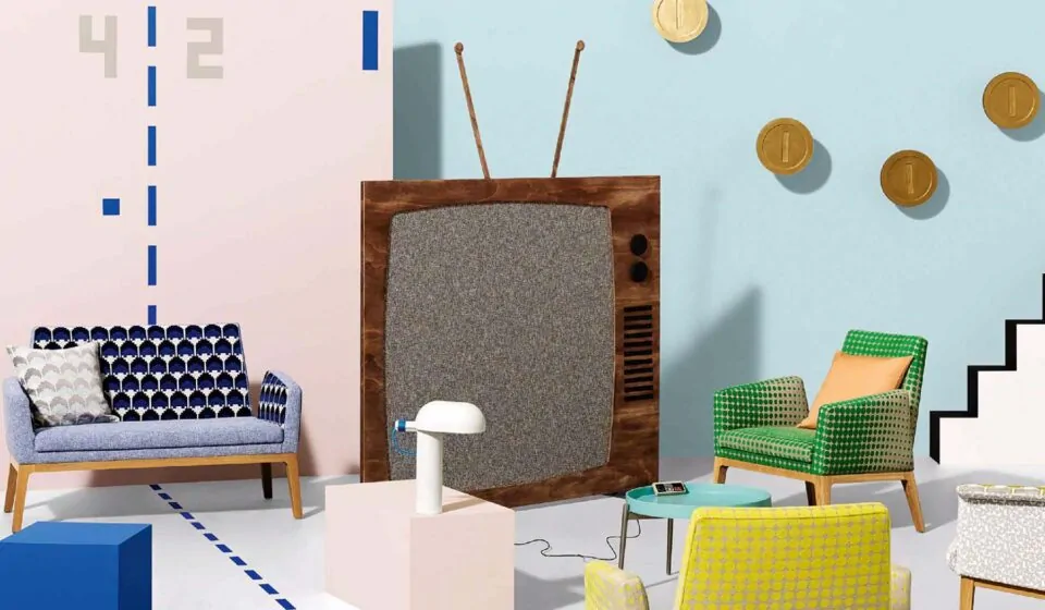 A vintage television stands prominently, connected to a modern lamp on a light-colored pedestal. Colorful chairs surround it, with graphic wall elements in a playful, bright room.