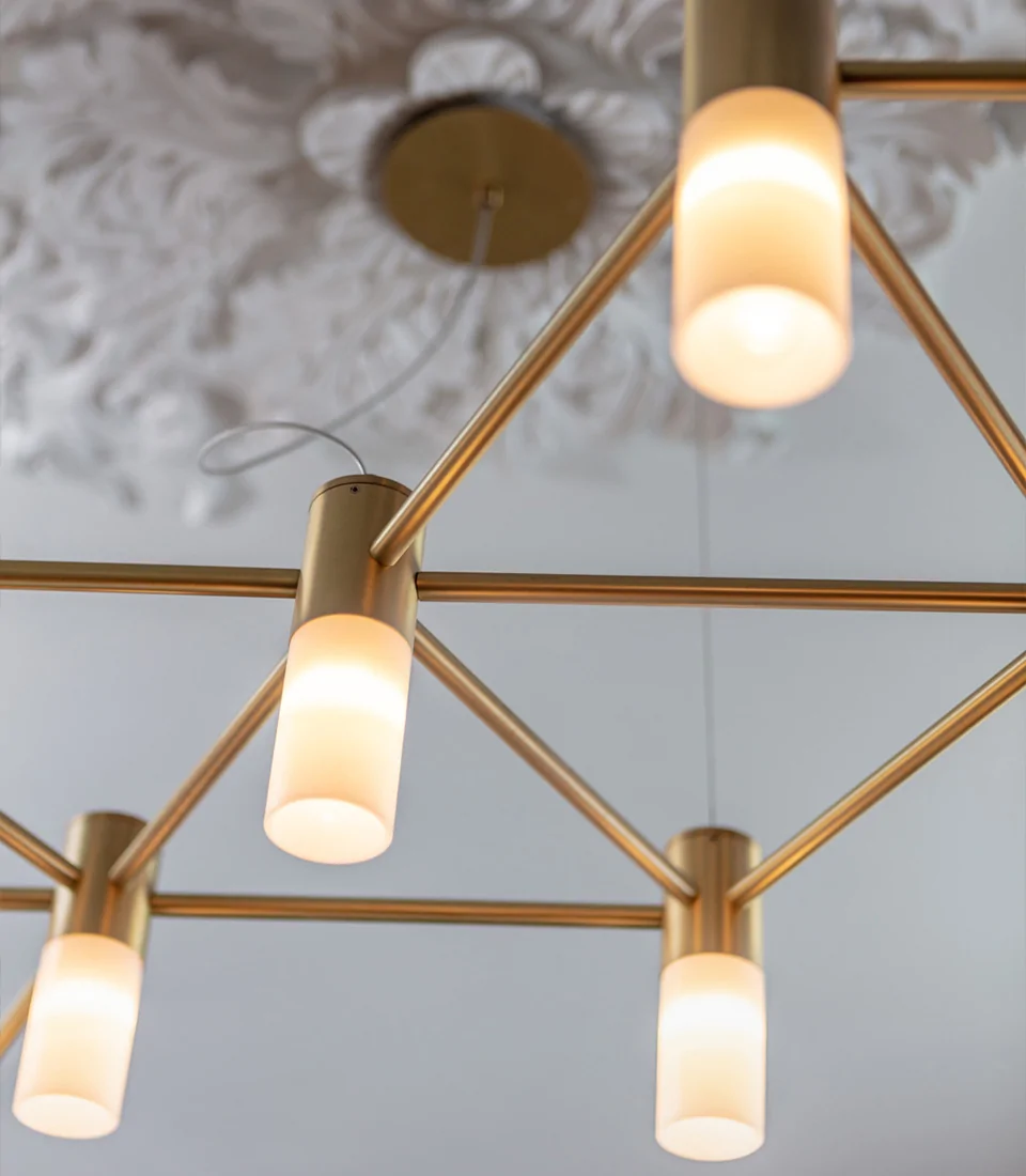 A modern ceiling lamp featuring multiple cylindrical light fixtures with frosted shades, suspended from a metallic bronze frame. The background shows an ornate white ceiling design.