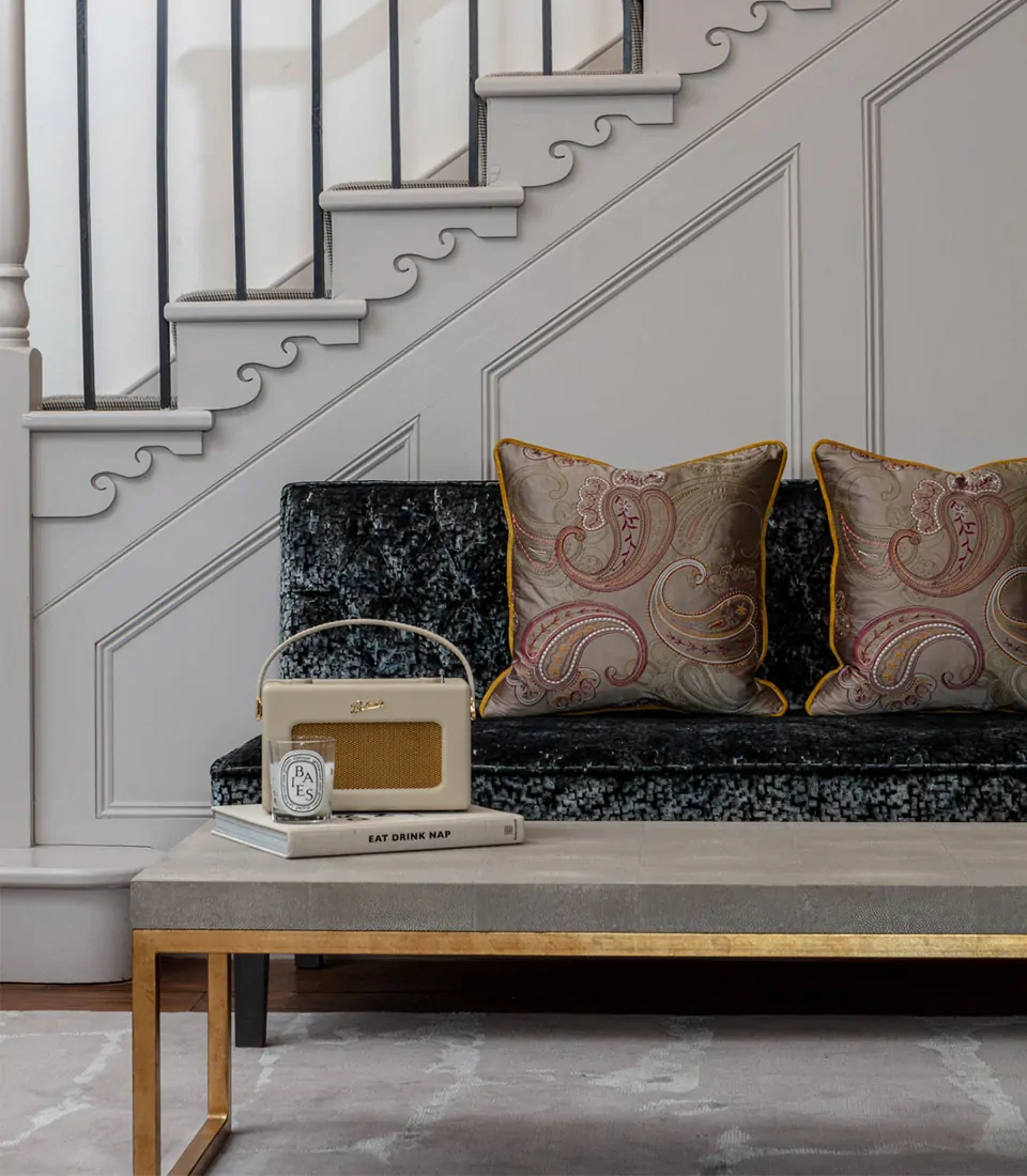 A decorative coffee table displays a small vintage radio and books. Behind it, a plush sofa with ornate cushions sits against a stairway, enhancing the cozy indoor ambiance.