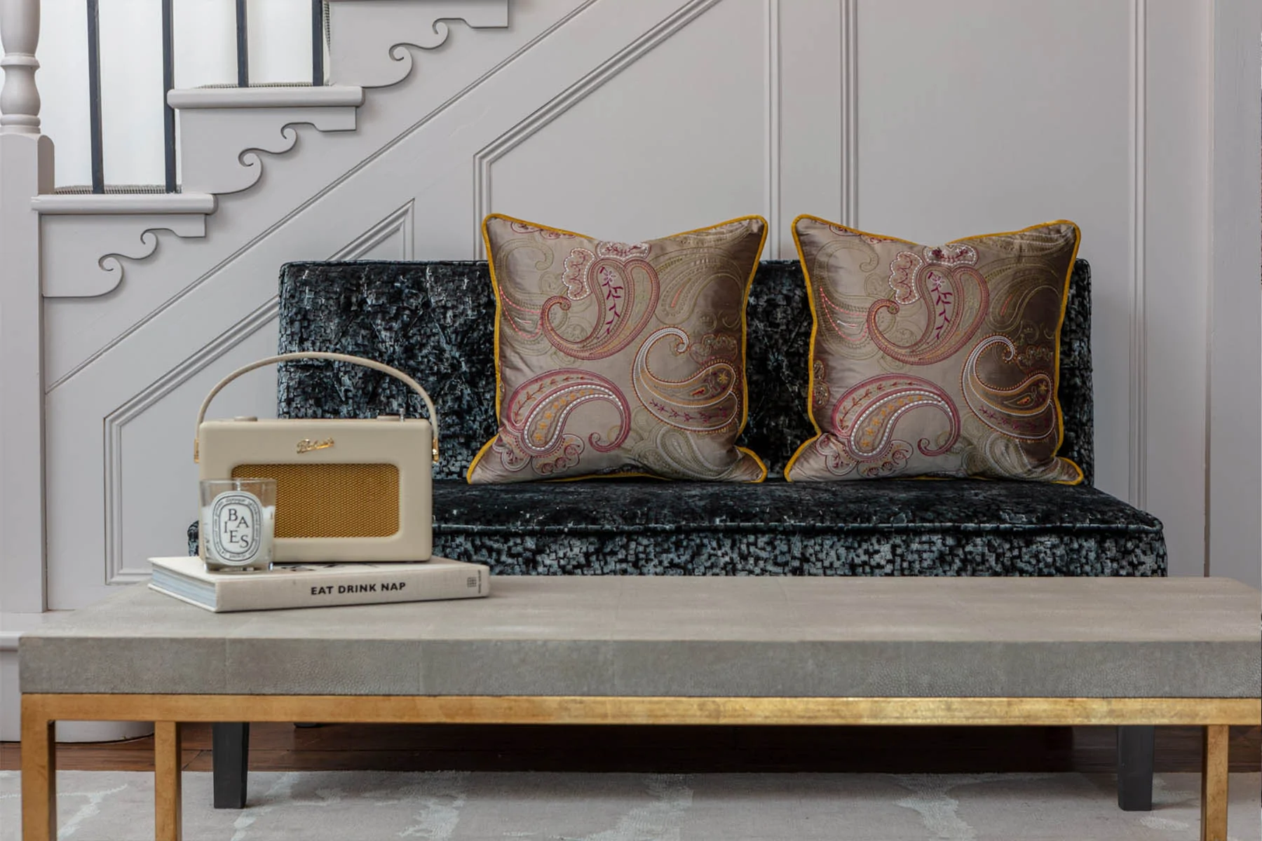 A plush, dark velvet sofa, adorned with two decorative paisley-patterned pillows, sits beside a small coffee table. A vintage radio and a candle rest atop the table, enhancing a cozy atmosphere.
