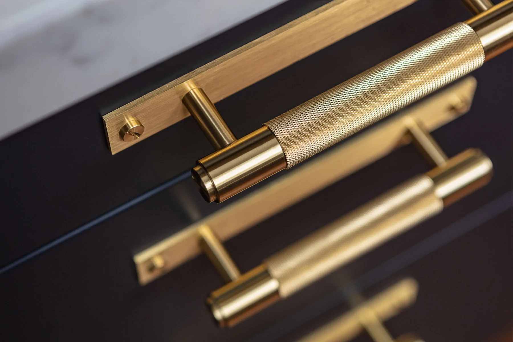 A polished brass cabinet handle is prominently displayed, featuring a textured grip. It's positioned against a dark cabinet surface, suggesting a contemporary design in a kitchen or furniture setting.