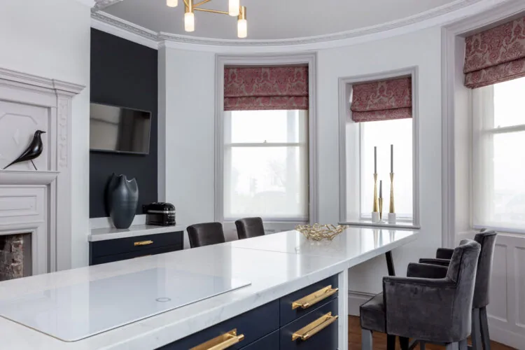 A modern kitchen features a marble-topped island, dark cabinetry, gold accents, and elegant decor elements. Large windows provide natural light, highlighting sleek furnishings and a stylish ceiling lamp.