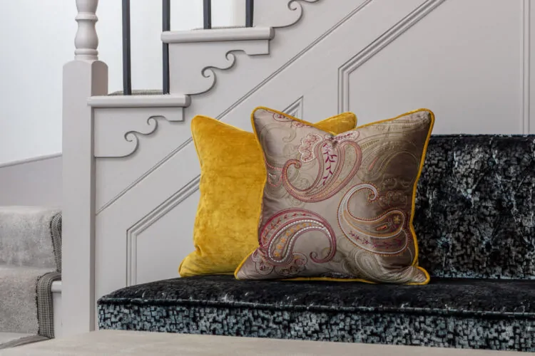 A pair of decorative pillows sit on a plush, dark upholstered sofa near a staircase. One pillow is vibrant yellow, while the other features an intricate paisley design, enhancing the stylish ambiance.