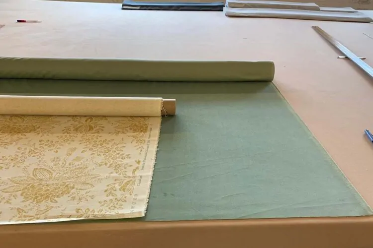 A roll of patterned fabric lies on a working table, partially unrolled next to a solid-colored green fabric. Various fabric rolls and tools are visible in the background.