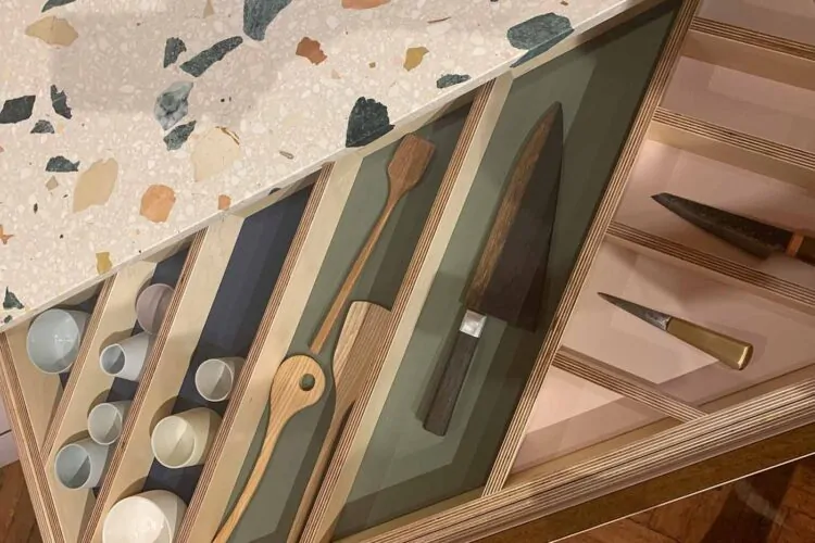 Wooden drawers display various kitchen utensils, including knives and stirring spoons. The countertop features a speckled design, adding a colorful, textured background to the organized layout of tools.