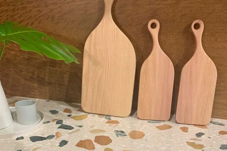 Three wooden serving boards of varying sizes stand upright against a brown wall. A green leaf plant and a small cup on a speckled countertop complete the kitchen setting.