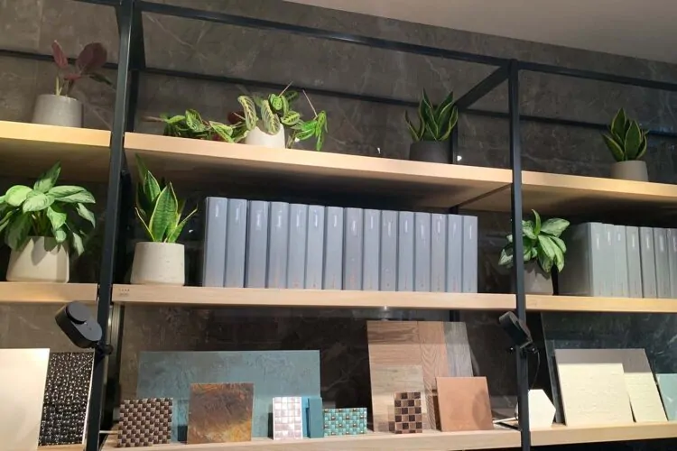 A shelf displays various potted plants alongside stacked gray binders and decorative samples, including tiles and wood pieces, against a marble wall in a modern indoor setting.
