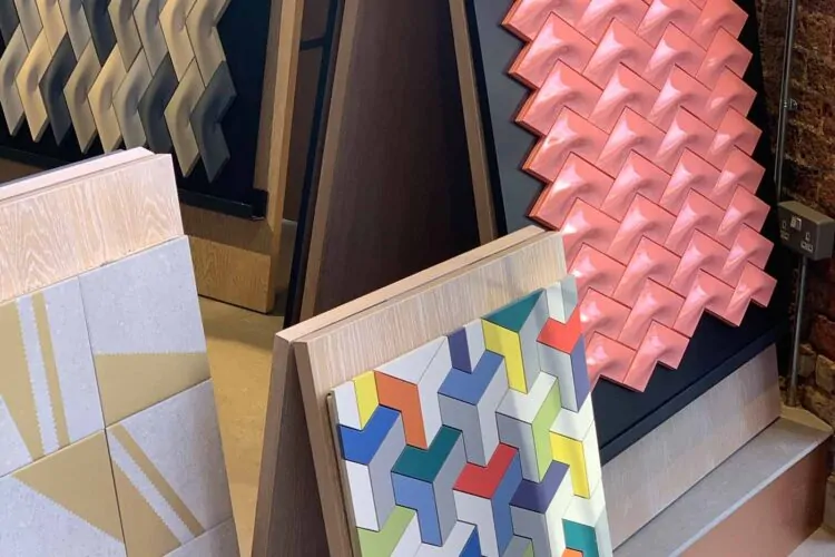 Colorful wall tiles in varying geometric patterns are displayed upright. They include textured designs in pink, gray, and multicolored shapes, set against a modern interior backdrop.