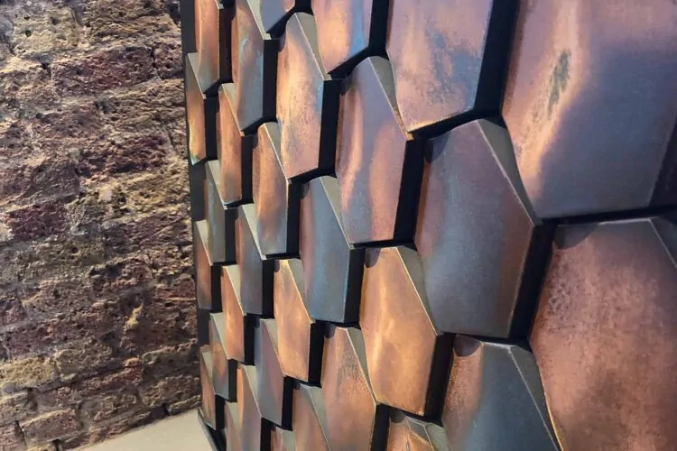 A hexagonal patterned wall surface showcases a blend of earthy tones, with textured elements catching light. Behind it, a rustic brick wall adds depth to the setting.
