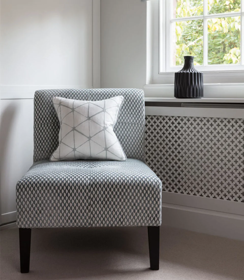 A patterned armchair with a decorative pillow sits beside a window, against a wall featuring intricate geometric designs. Light filters through, creating a calm, inviting atmosphere.