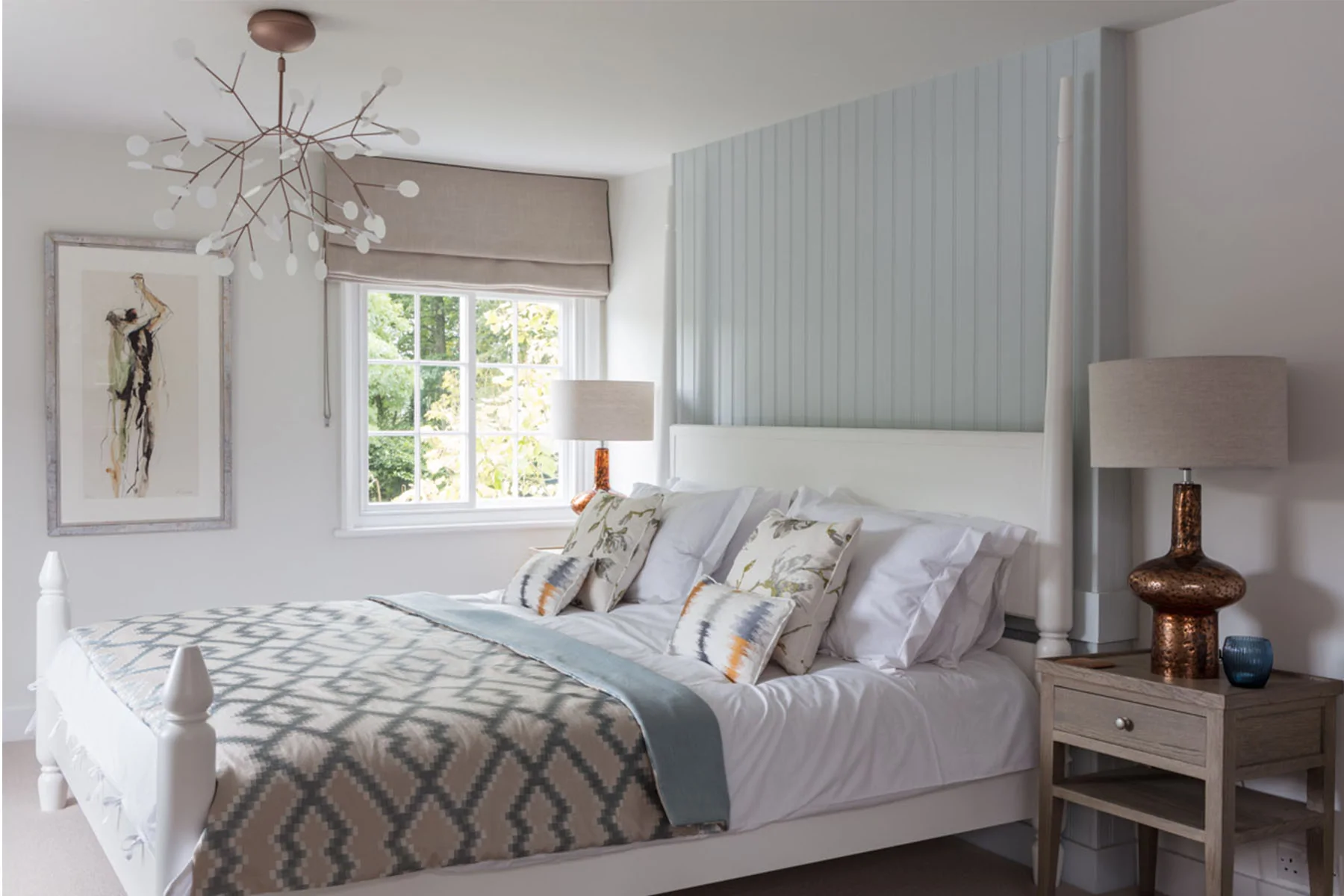A double bed with decorative pillows is neatly arranged in a bright bedroom. A stylish chandelier hangs above, while natural light filters through a window beside a bedside table with a lamp.
