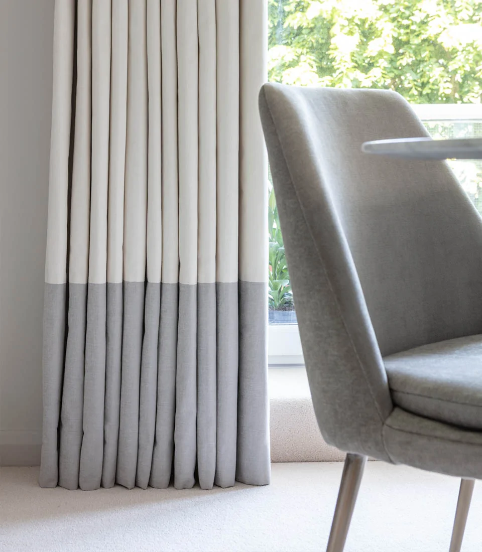 A set of elegant, pleated curtains in white and gray drapes by a window, while a modern gray chair sits next to a small, light-colored table, with green foliage visible outside.