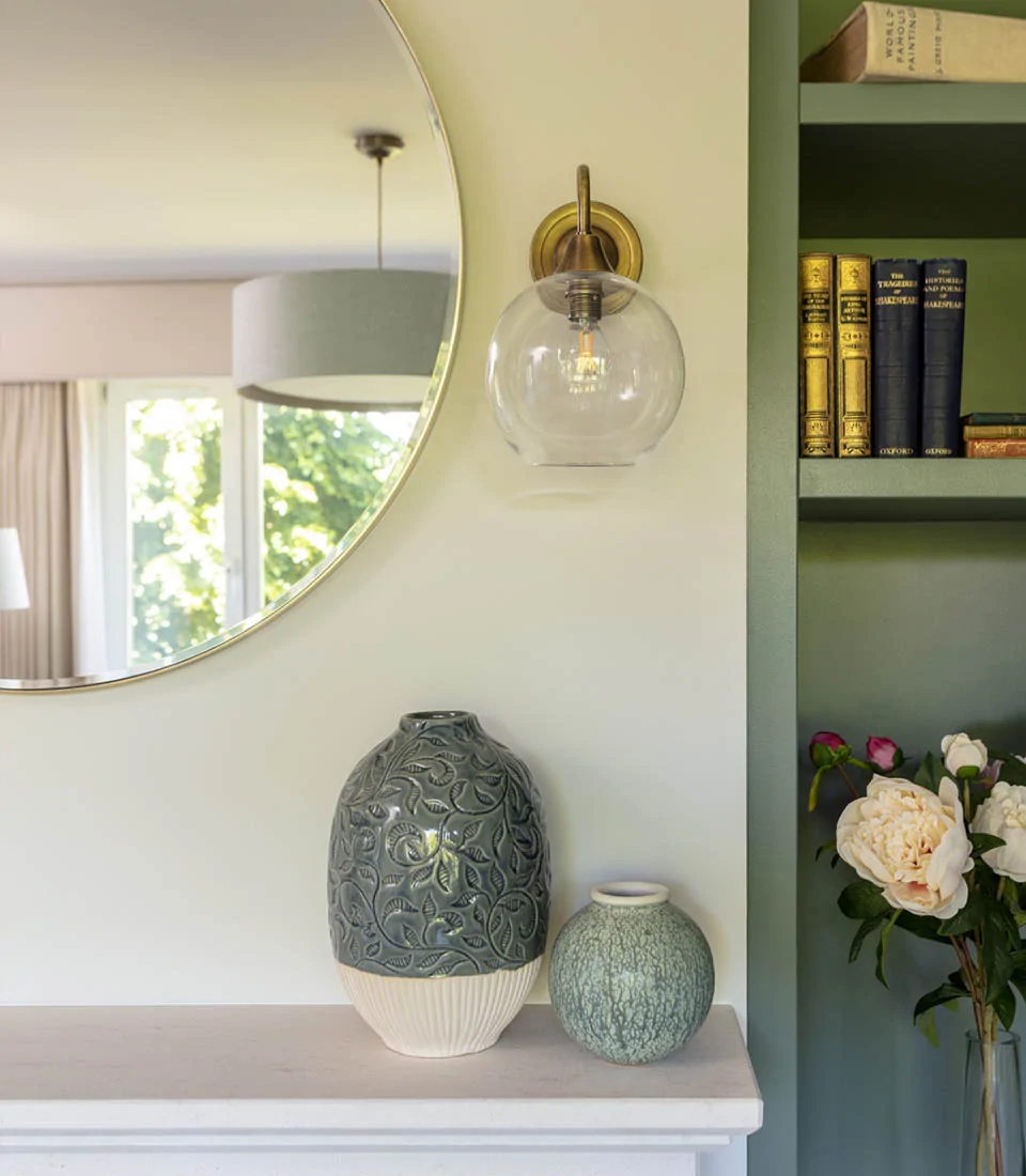 A decorative mirror reflects a clear glass wall light above two intricate ceramic vases on a mantel. A nearby bookshelf holds various books, and a bouquet of peonies adds color.