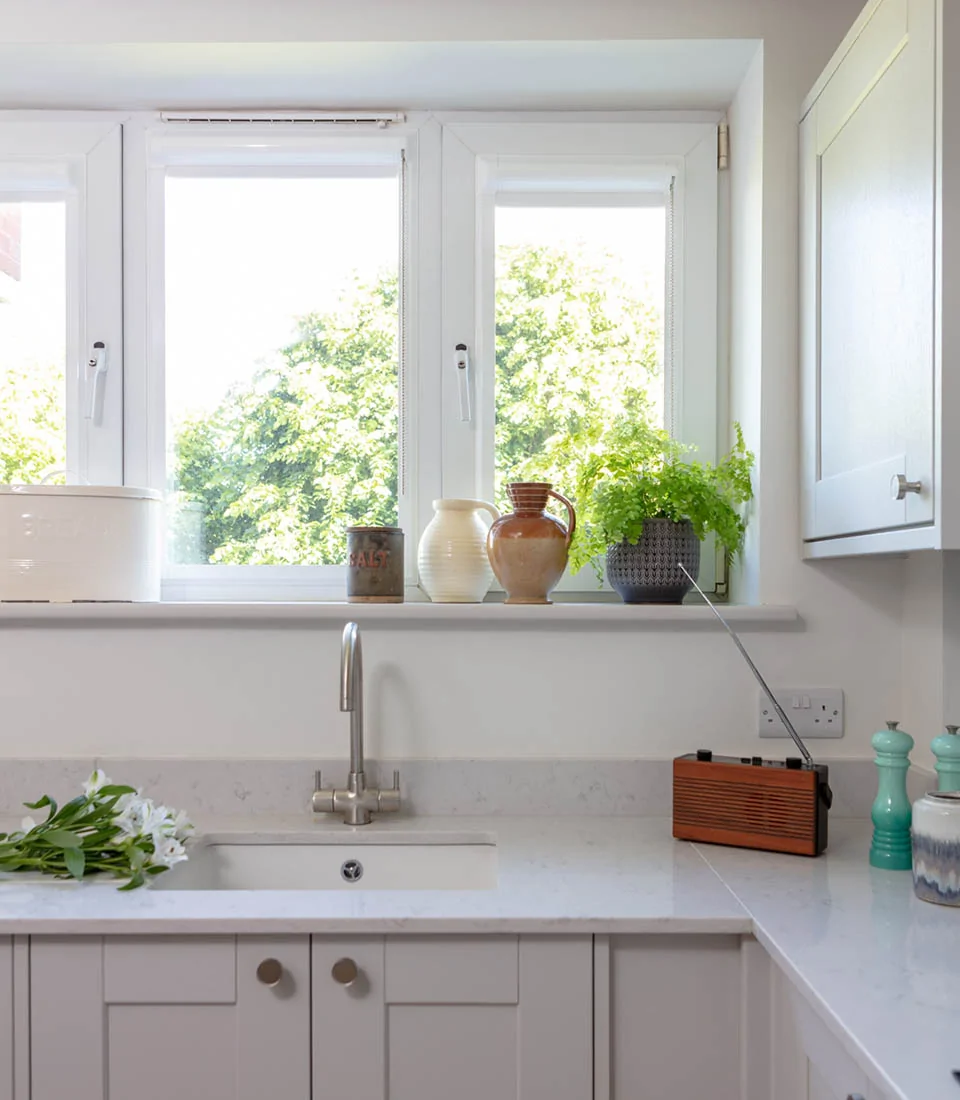 A modern kitchen features a sink and countertop with fresh flowers. Sunlight streams through windows, illuminating decorative pots and an antique radio on the windowsill, against a backdrop of greenery.