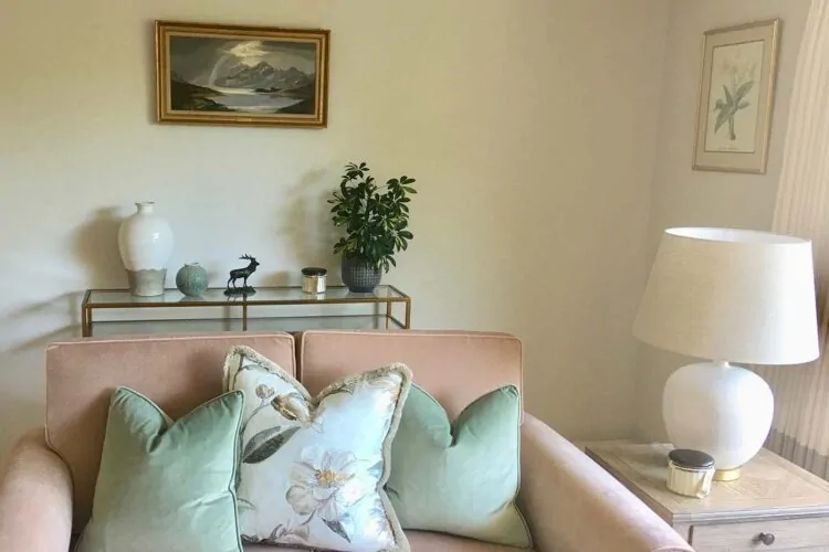 A soft pink sofa adorned with decorative pillows sits in a bright room. A glass console displays vases and a small plant, while a nearby lamp illuminates the space. A landscape painting hangs above.