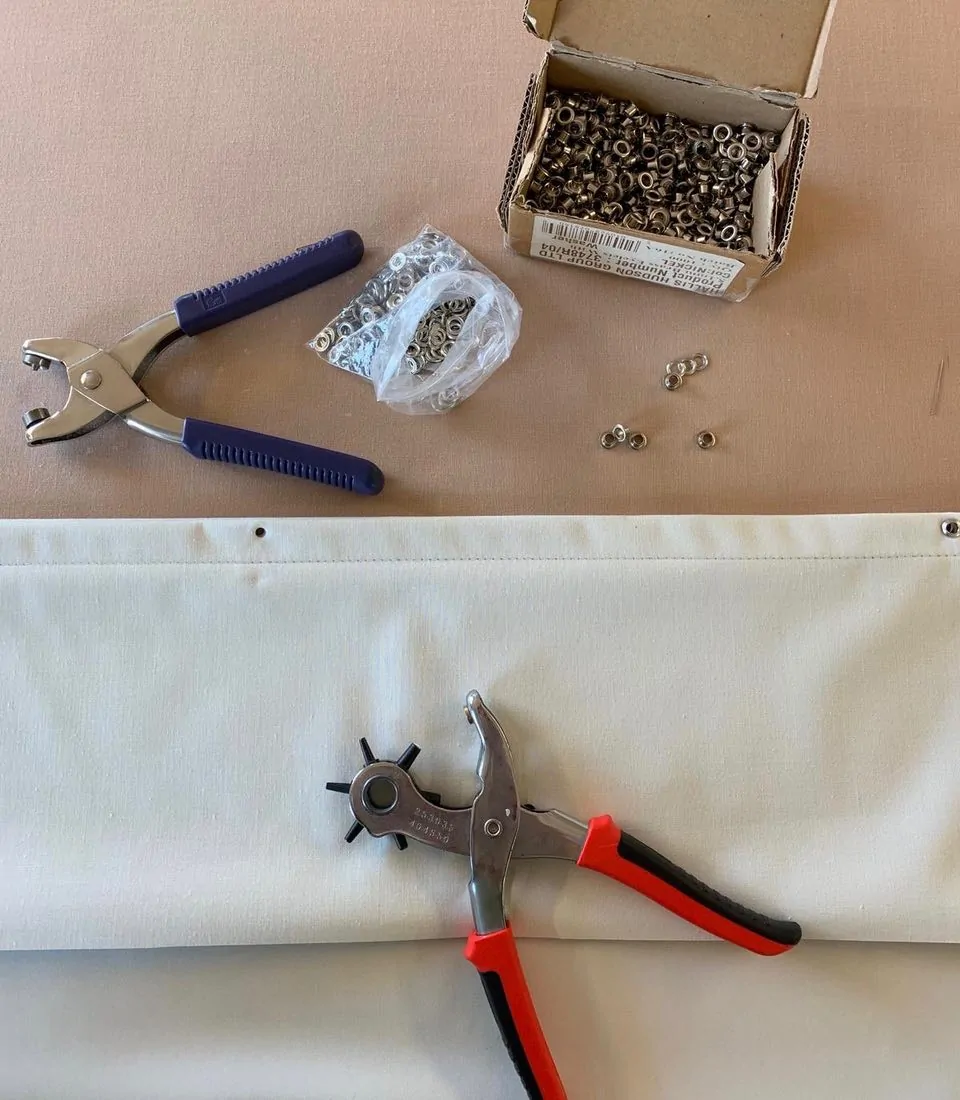 A pair of grommet pliers is positioned next to a piece of fabric with a hole, surrounded by a box and a bag of metal grommets on a beige surface.