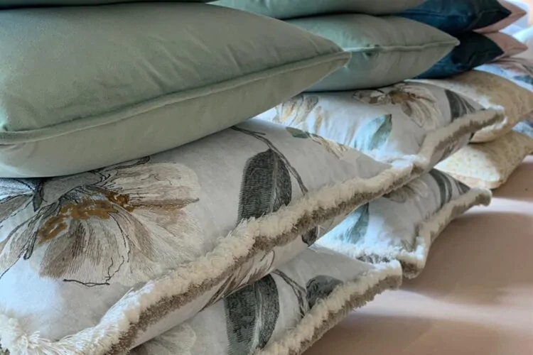 A stack of decorative cushions is arranged neatly. The top cushions are solid green, while lower ones feature floral patterns with fringe edges. They rest on a light-colored surface.