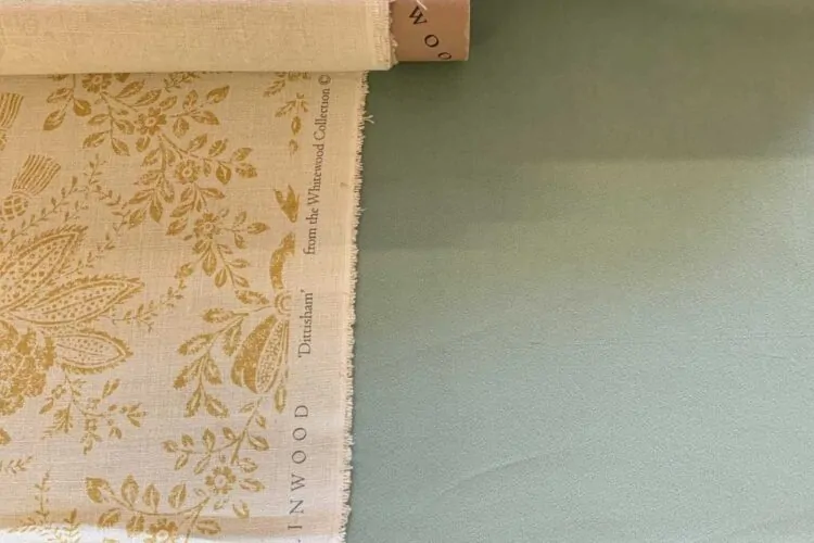 A patterned fabric roll, featuring yellow floral designs on a light background, is partially unrolled on a smooth, muted green surface. The fabric is labeled "LINWOOD" and "from the Whitewood Collection."